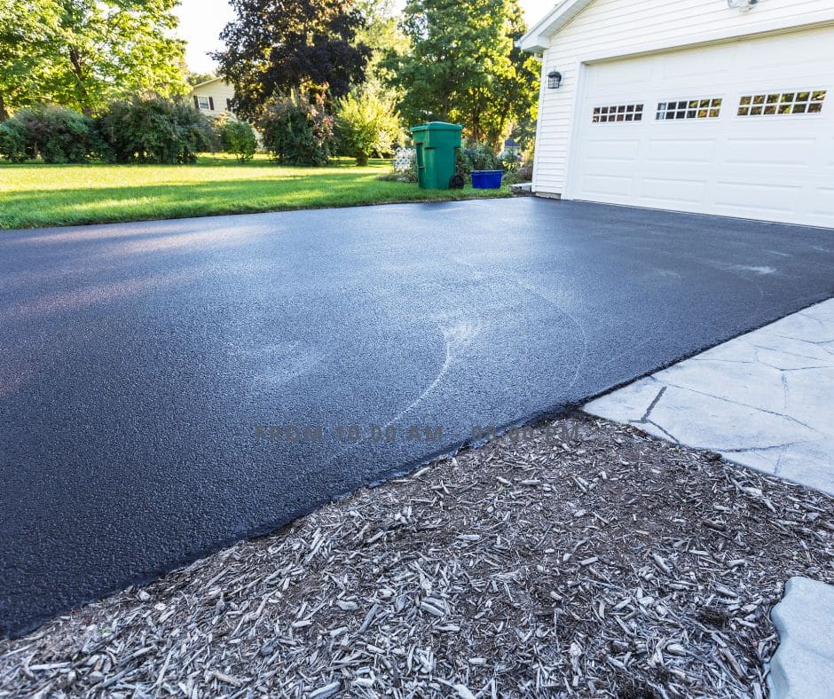 Newly resurfaced driveway leading to a white garage door, bordered by mulch and green lawn, in a suburban setting.