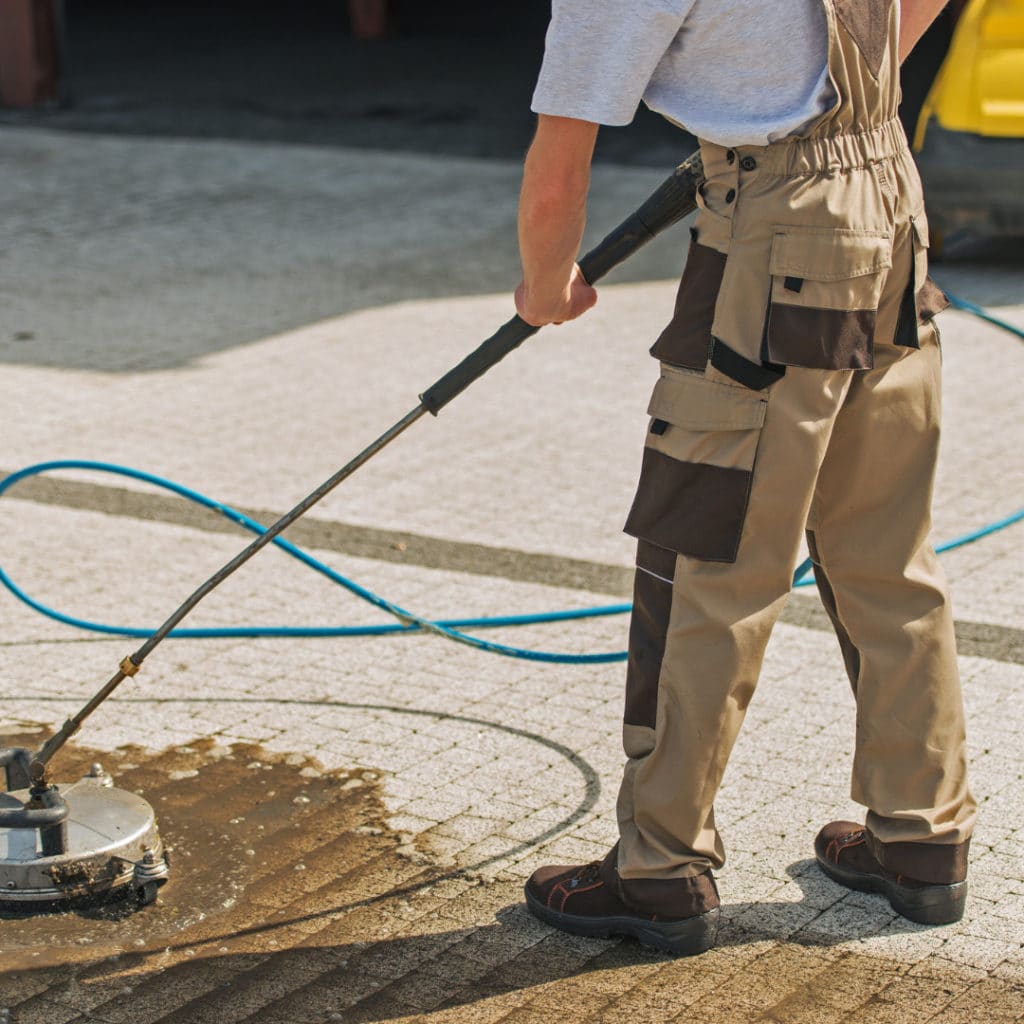 Worker in beige overalls using a high-pressure washer on a concrete surface with a blue hose trailing behind.