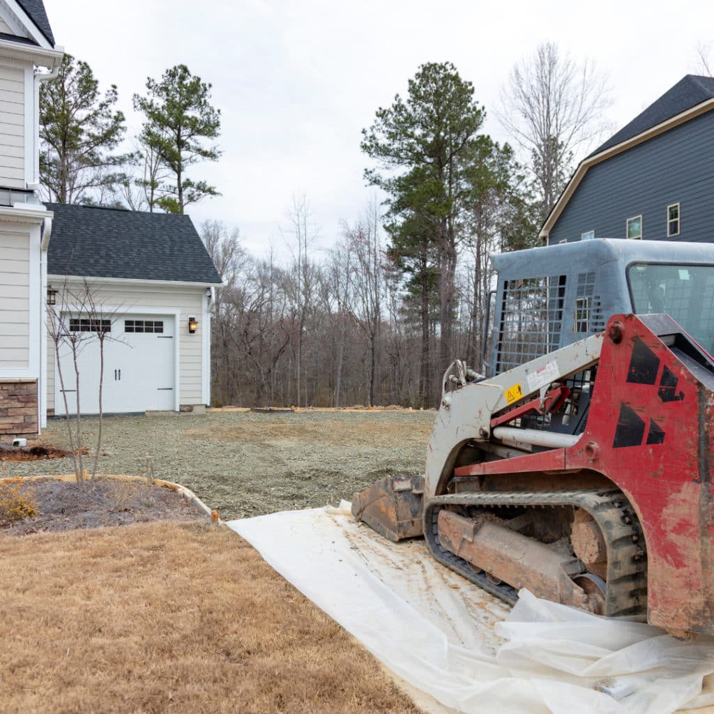 Residential construction site with red skid steer loader on gravel beside a new house with white garage door and bare trees in the background.