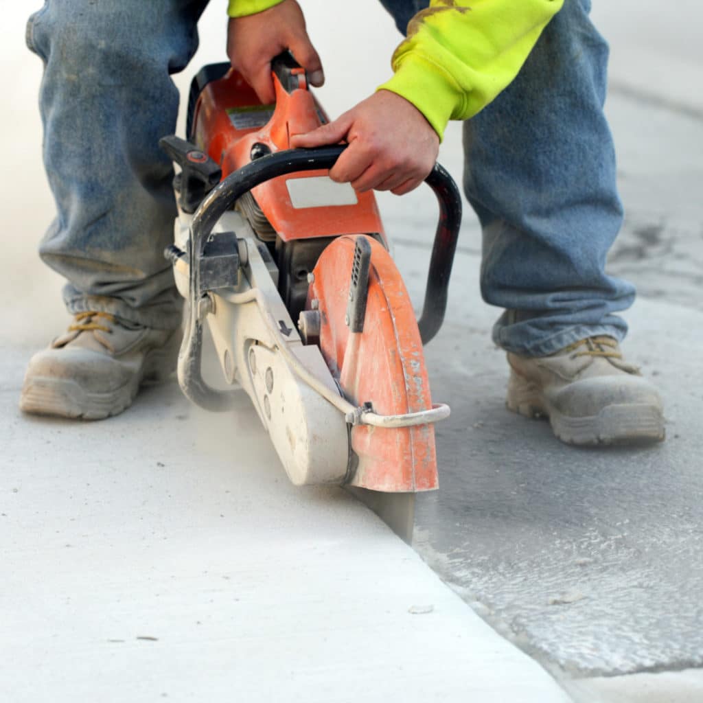 Worker in a high-visibility jacket using an orange concrete saw to cut through a gray concrete surface, generating dust, wearing safety boots.
