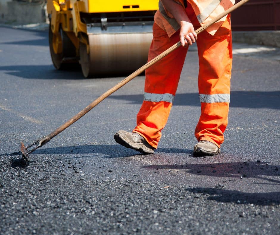 Close-up of a construction worker in orange safety pants using a rake to spread asphalt with a steamroller in the background.