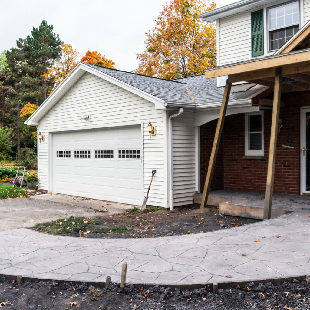 A residential driveway with freshly laid patterned concrete leading to a white garage, surrounded by autumn foliage.