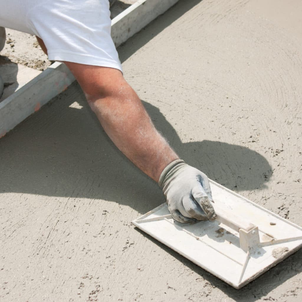 Close-up of a worker's hand using a trowel to smooth wet concrete with wooden forms in the background.