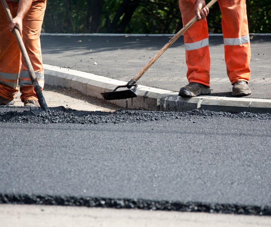 Road workers in high-visibility clothing using rakes to spread hot asphalt next to a curb on a sunny day.