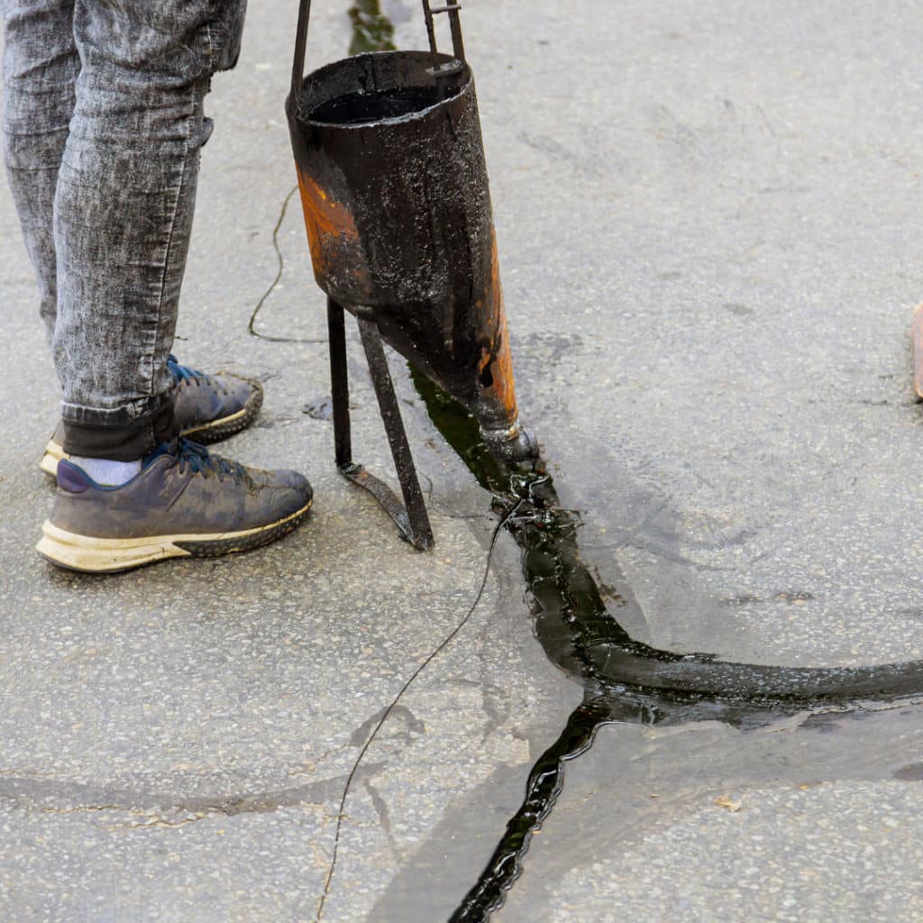 Worker pouring tar to seal a crack in the asphalt, with a focus on the pouring bucket and the black sealant.