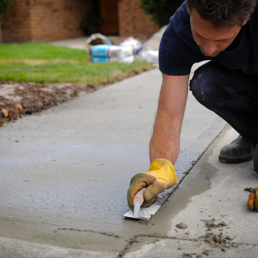 A worker in a crouched position smoothing concrete with a trowel on a driveway, with construction materials in the background.
