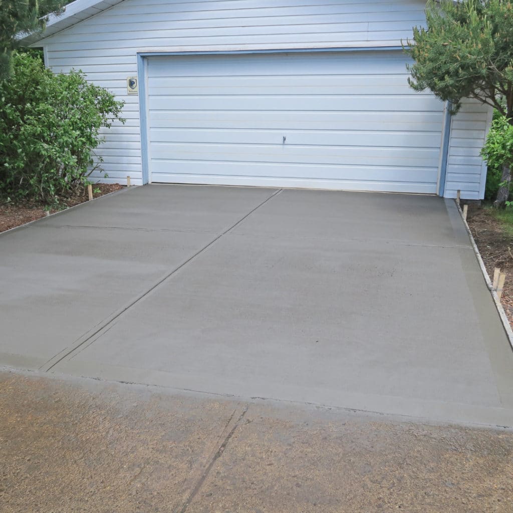 Freshly poured concrete driveway leading to a closed garage door with a neatly trimmed hedge on the left side.