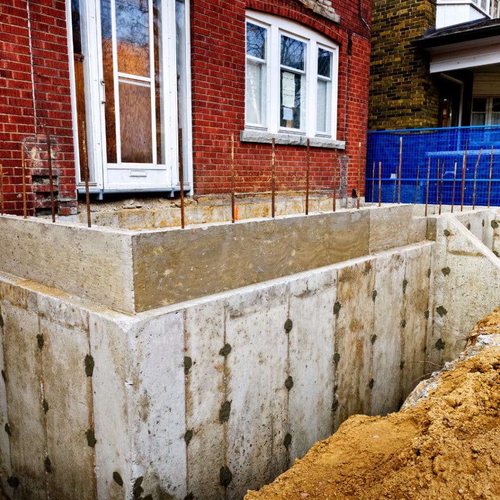 Home renovation showing a new concrete foundation extension in front of a red brick house with a blue safety fence.