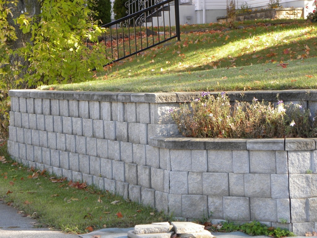Textured gray block retaining wall with overhanging greenery and a wrought iron fence on a sunny day.