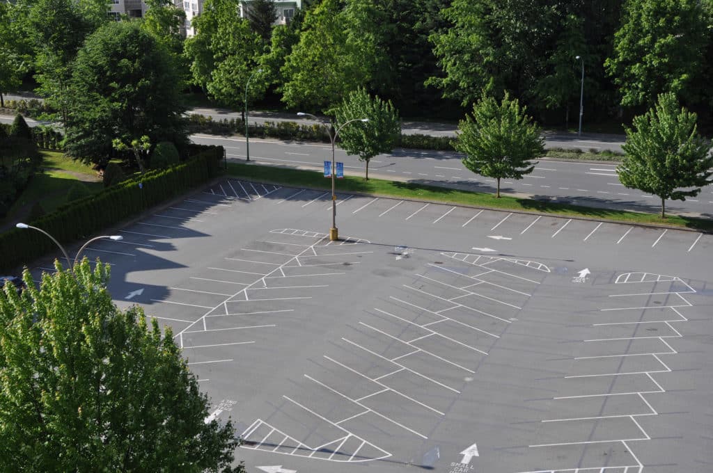 Aerial view of an empty parking lot with white lines and directional arrows, surrounded by green trees and hedges.