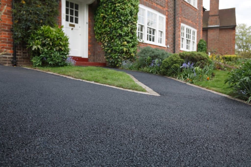 Freshly paved black driveway leading to a white door of a brick house with lush greenery and a garden.