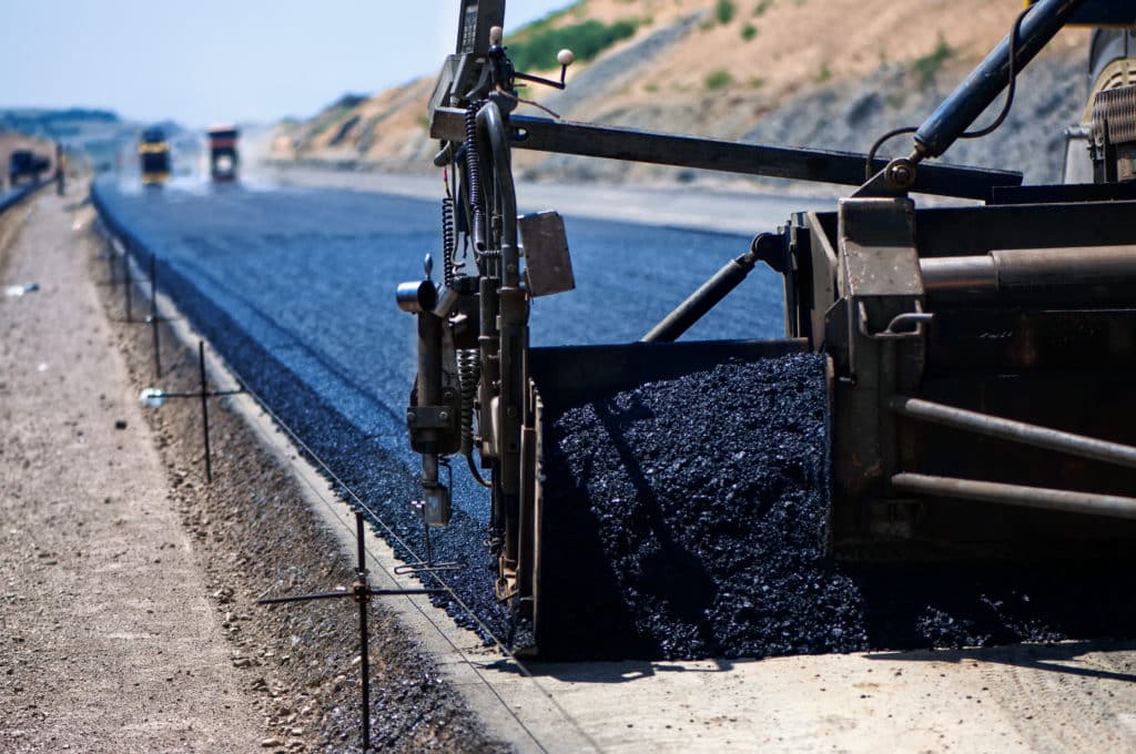 Close-up of an asphalt paver machine laying a new road surface, with construction vehicles in the distance.