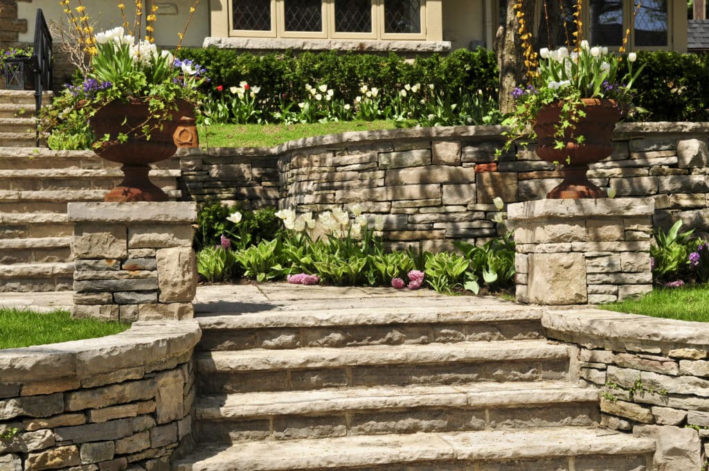 Stone steps leading to a landscaped garden with colorful flowers and ornamental pots in front of a residential home.