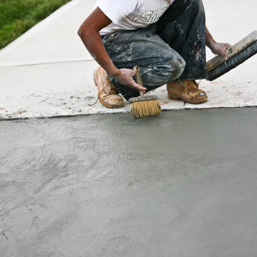 Close-up of a construction worker applying a textured finish to wet concrete with a hand brush.