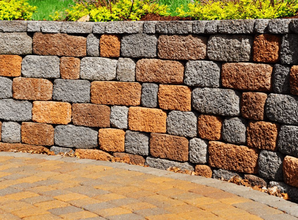 Textured multicolored paving stones forming a patterned retaining wall, with greenery above and matching pavement below.