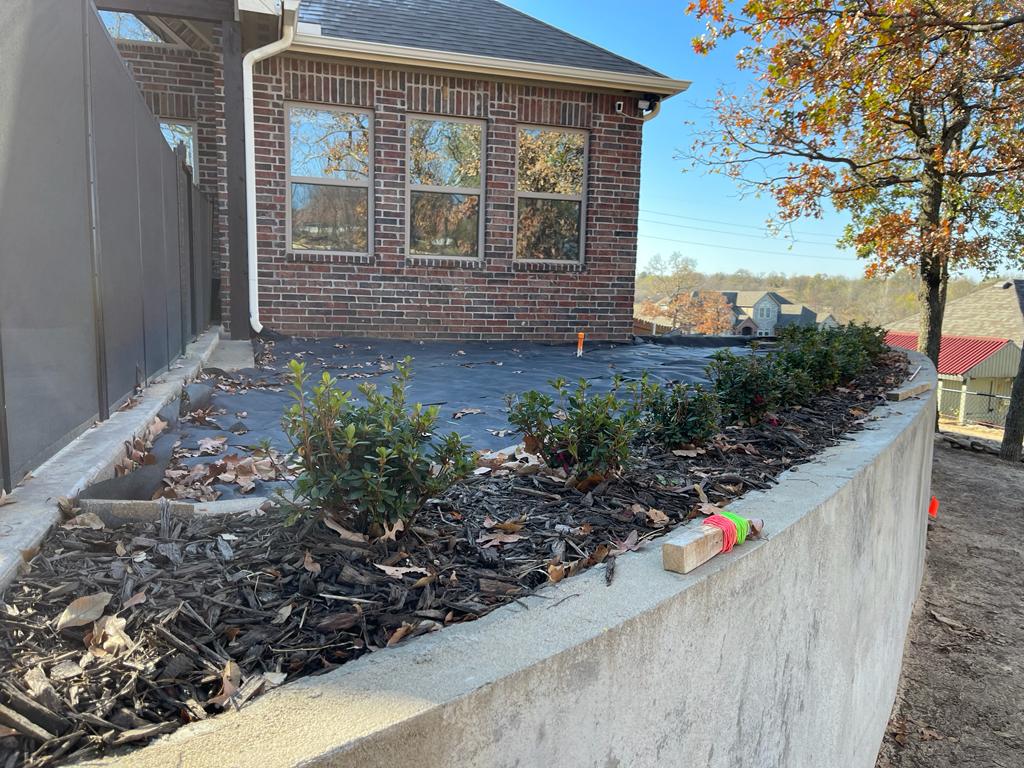Freshly mulched garden bed with young shrubs alongside a brick house with a window, under autumn trees.