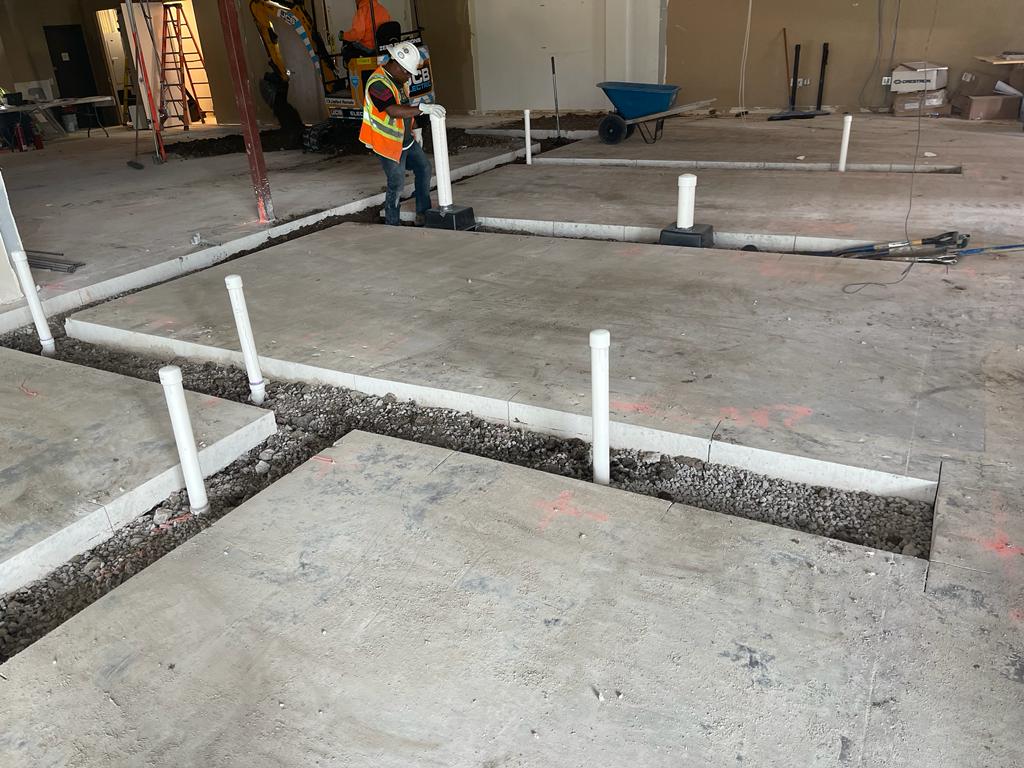 A construction site with a freshly poured concrete foundation and floor with several PVC plumbing stub-outs, a wheelbarrow, and a worker in the background.