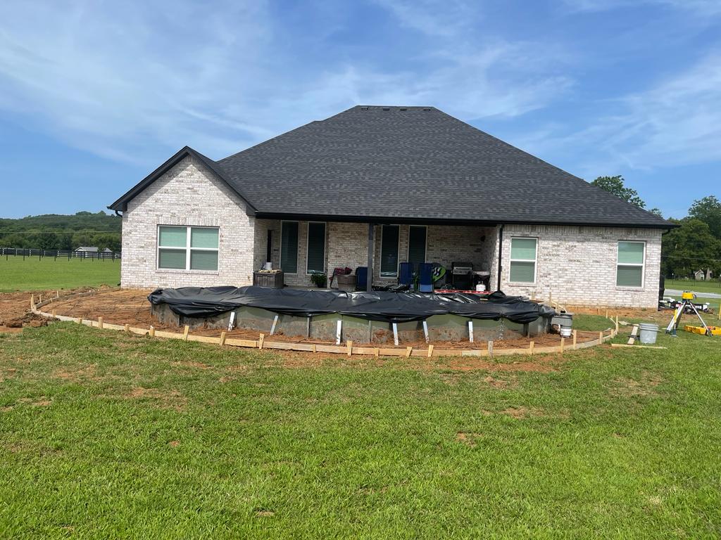 Brick house with a large backyard in the process of getting a new circular patio, covered with a black tarp and surrounded by lush green grass.