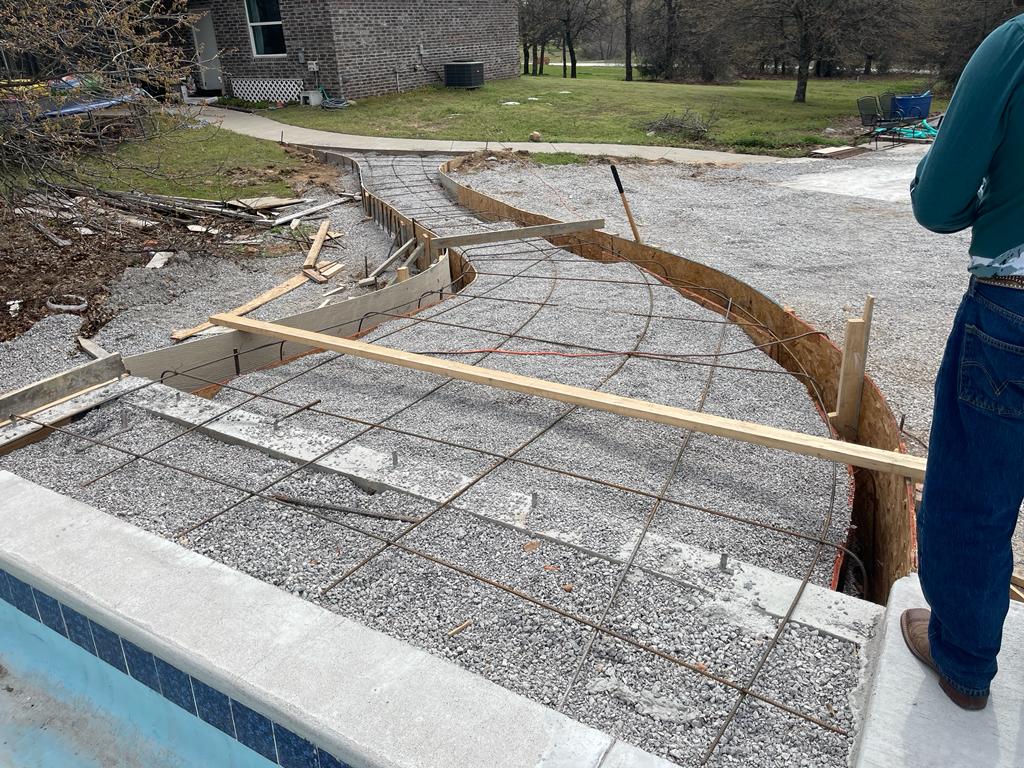 In-progress installation of a concrete patio featuring a rebar grid within wooden forms, next to a poolside in a residential area.