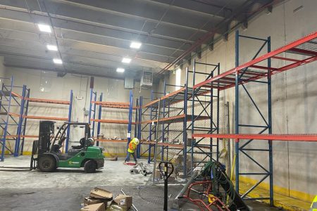 An indoor industrial site with a worker in a high-visibility jacket, storage racks, a forklift, and various construction equipment.